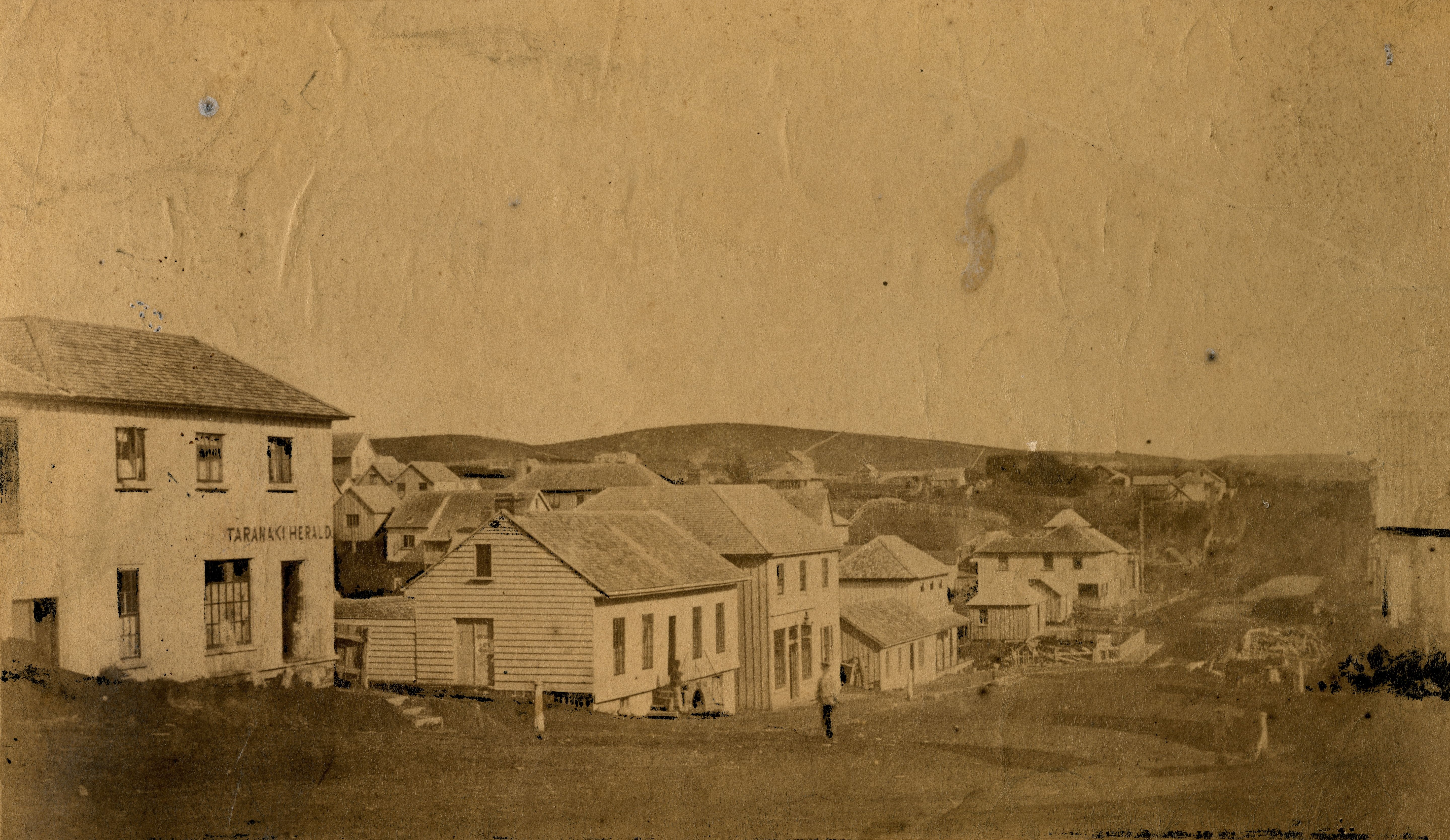 South side of Devon Street, New Plymouth with the original Taranaki Herald building on the left (1859). Hartley Webster. Collection of Puke Ariki (PHO2004-182).